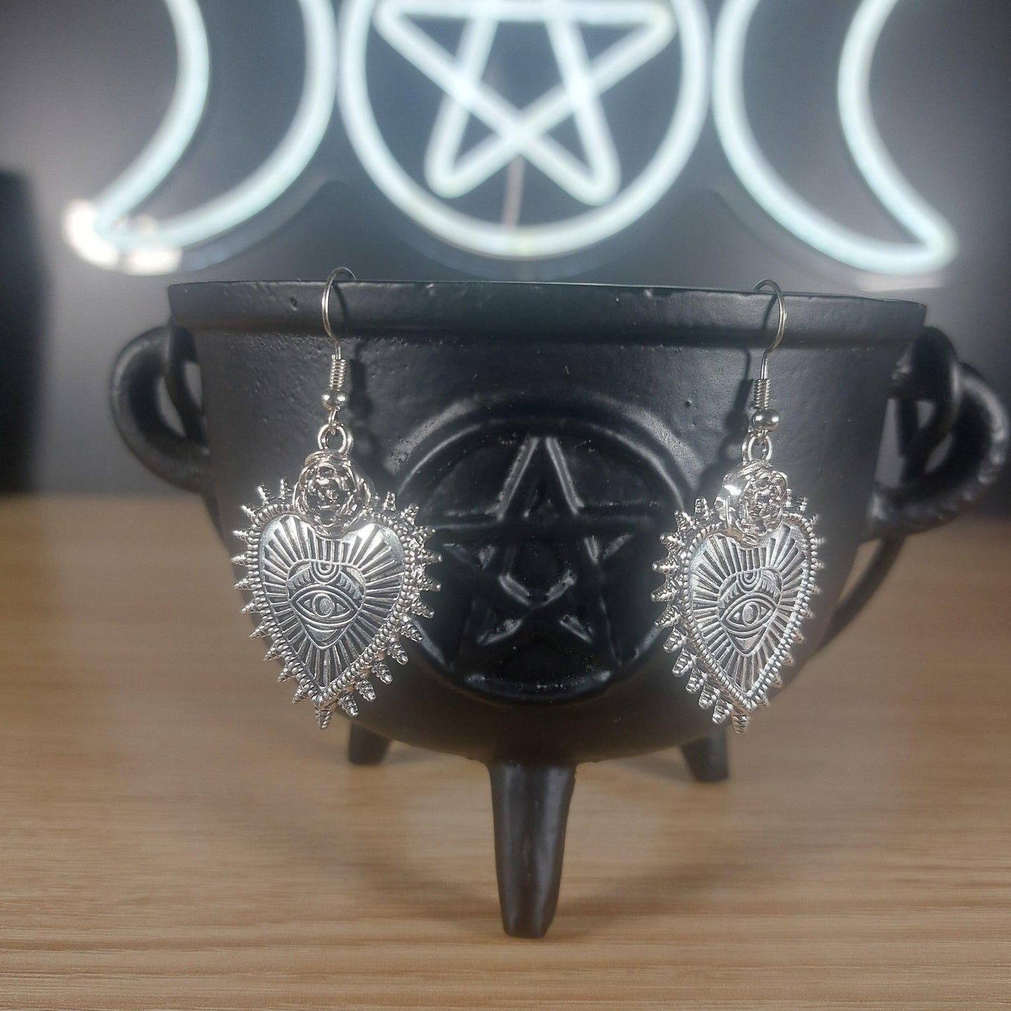Silver evil eye heart dangle earrings hanging from a black cauldron, with a triple moon and pentagram neon sign in the background.