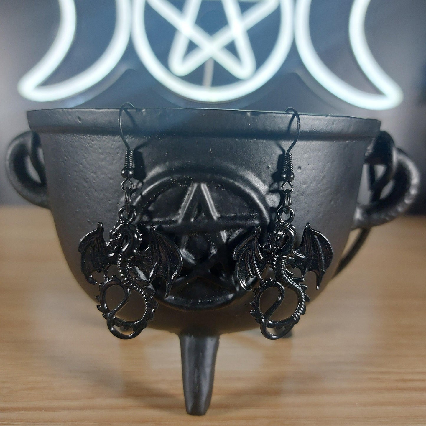 Black metal dragon dangle earrings with intricate wings, hanging from a black cauldron.