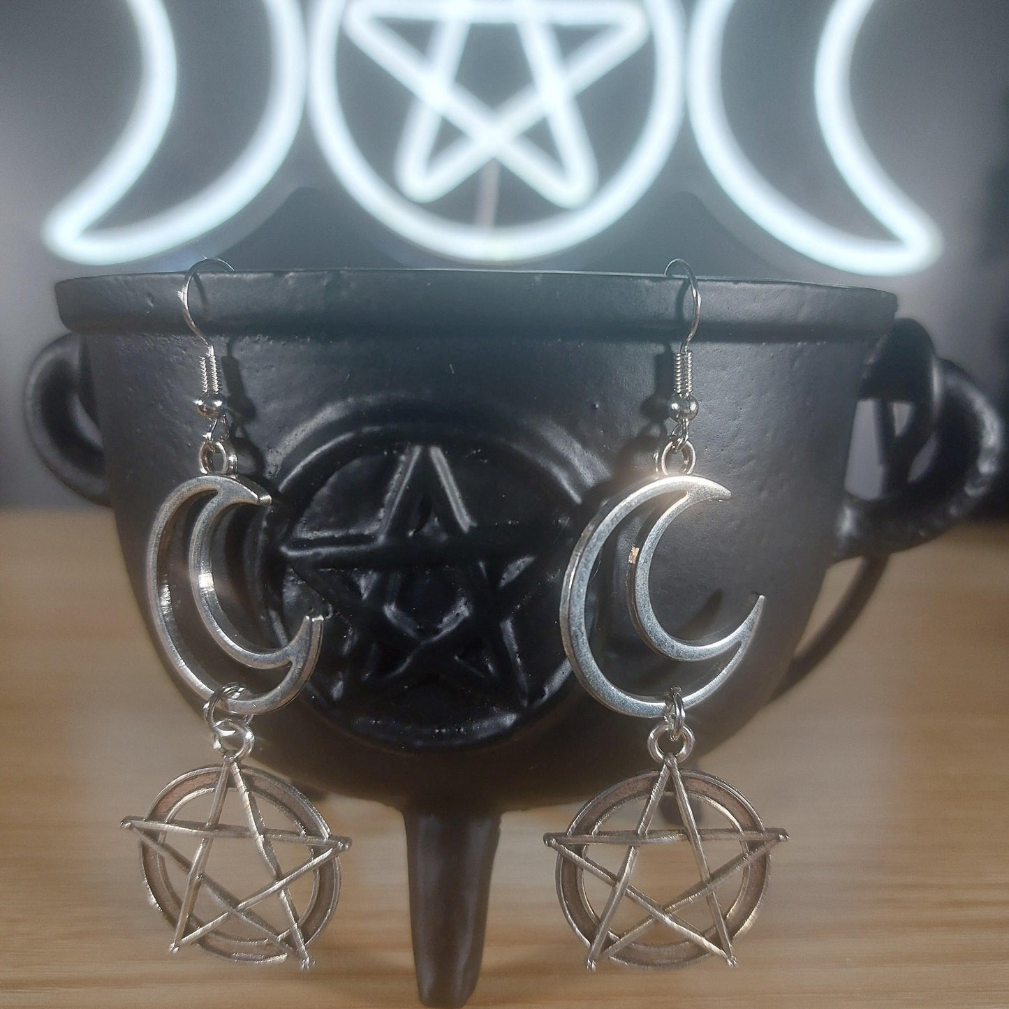 Silver moon and pentagram dangle earrings on a black cauldron, with a glowing triple moon symbol in the background.