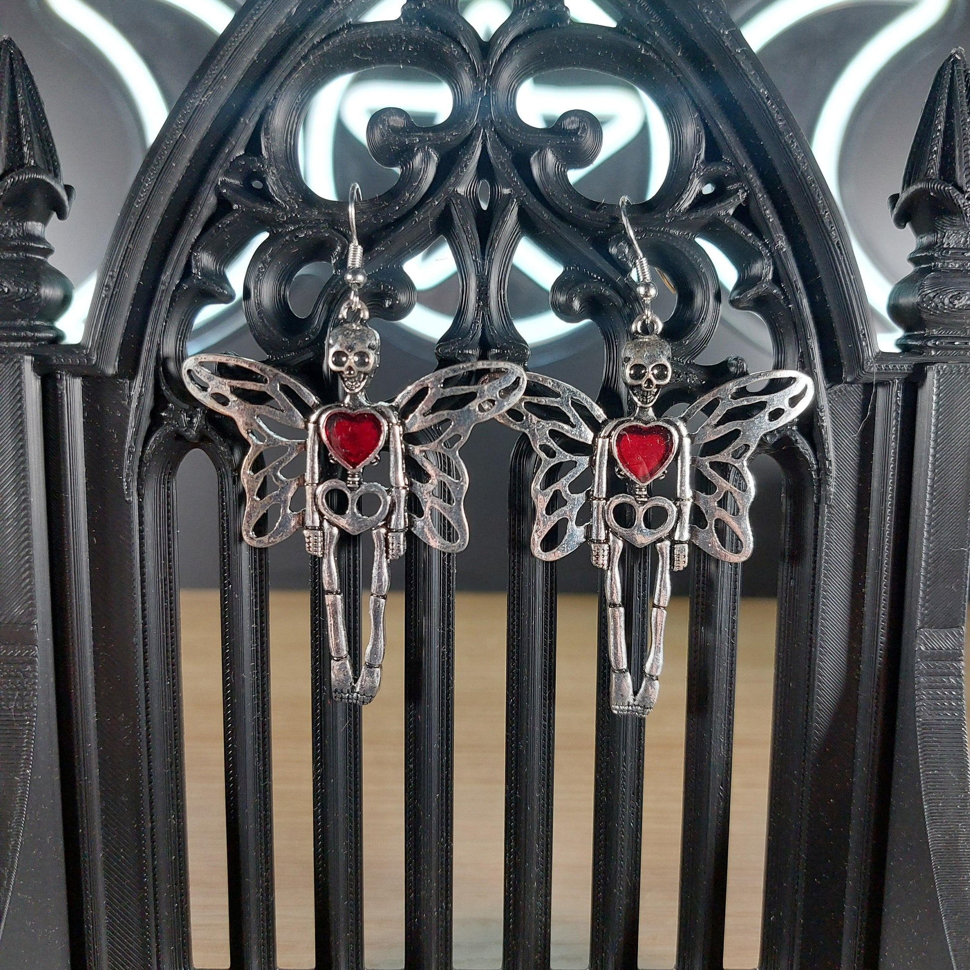 Silver tone skeleton dangle earrings with wings and a red heart, displayed on a black gothic arch.