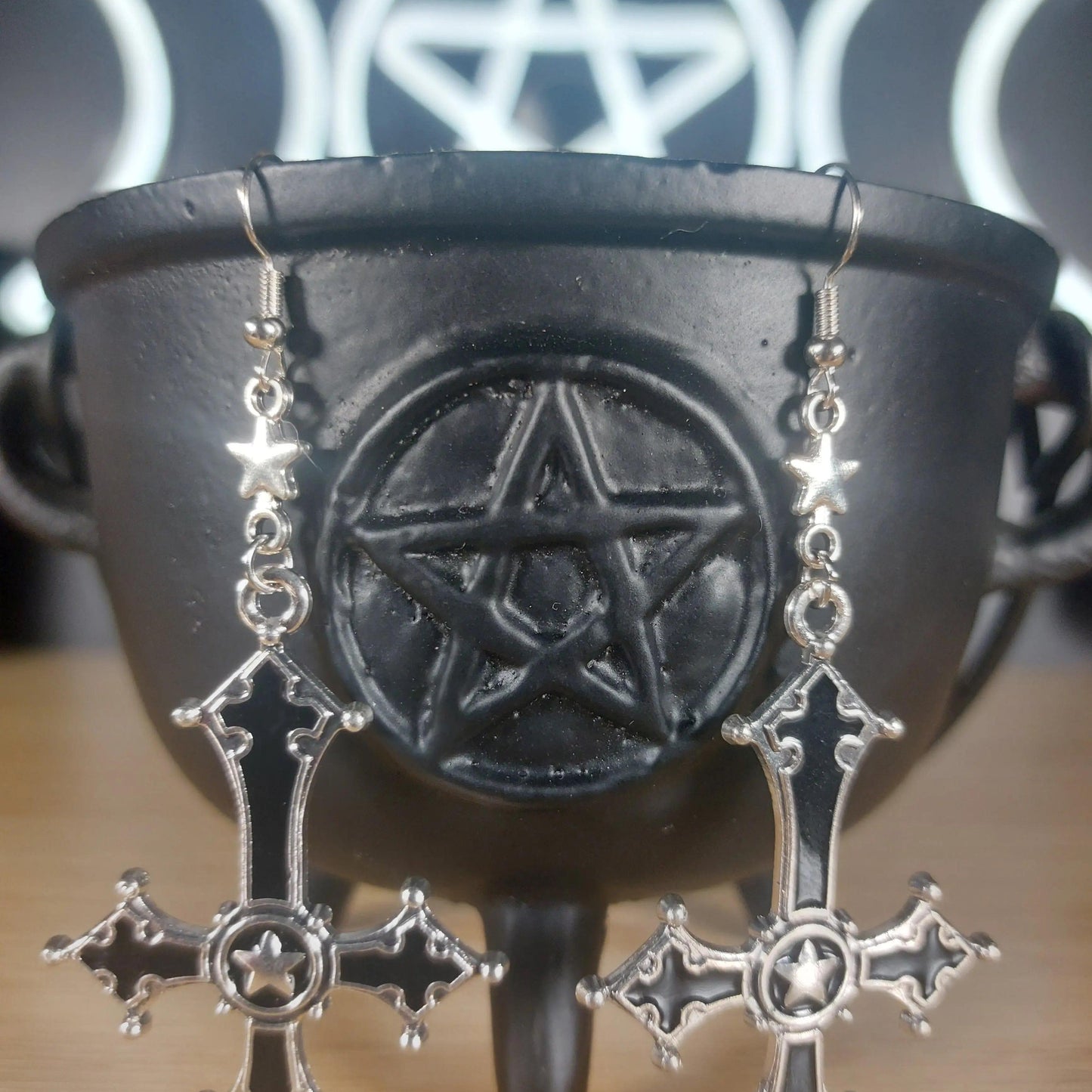 Silver and black upside-down cross earrings with star accents, hanging from a black cauldron.