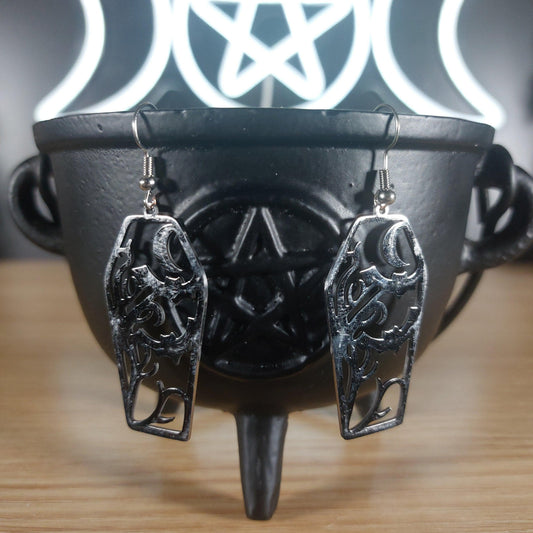 Silver tone coffin dangle earrings with cutout bats and moon design, hanging from a black cauldron.