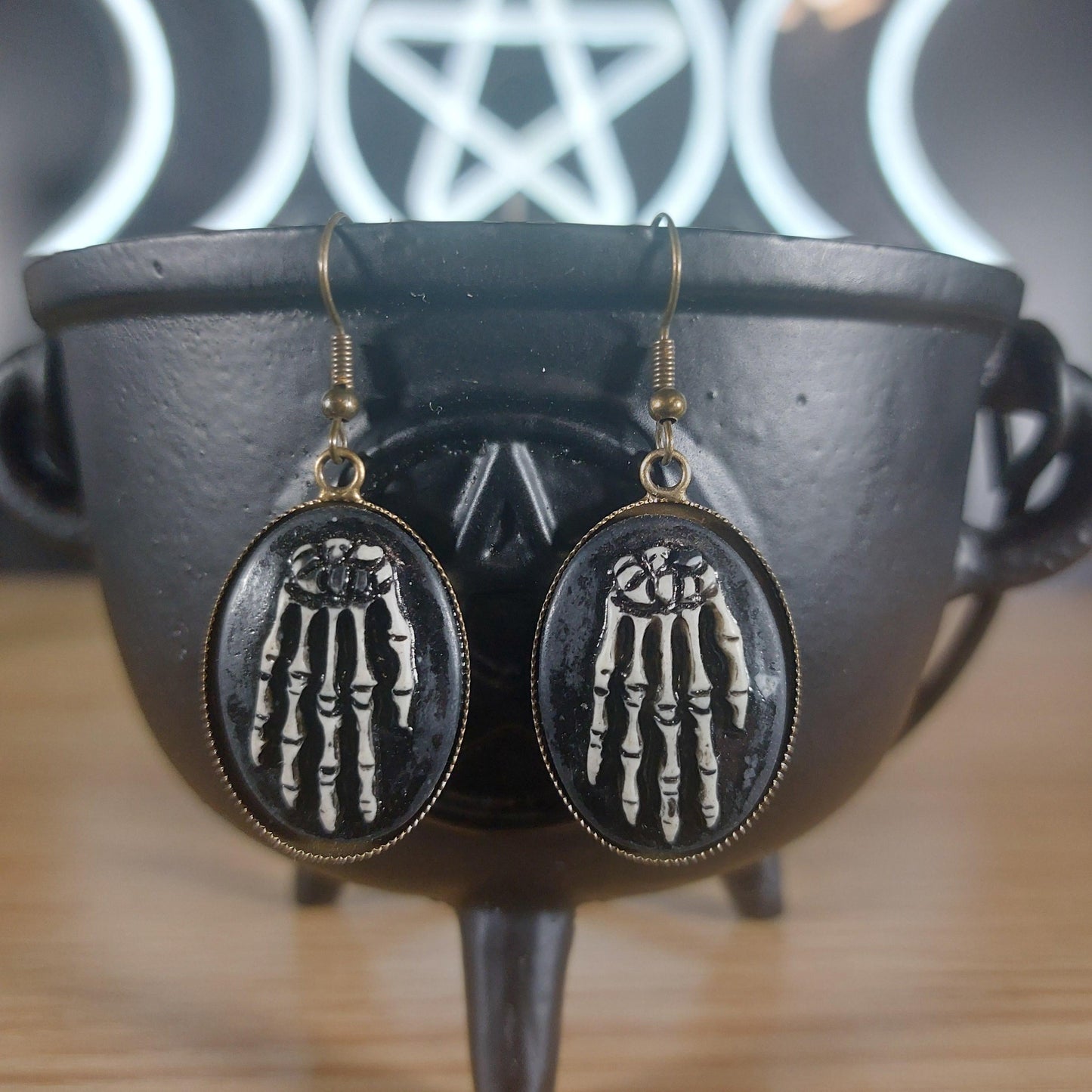 Antique bronze skeleton hands dangle earrings with black oval frames, resting on a black cauldron.