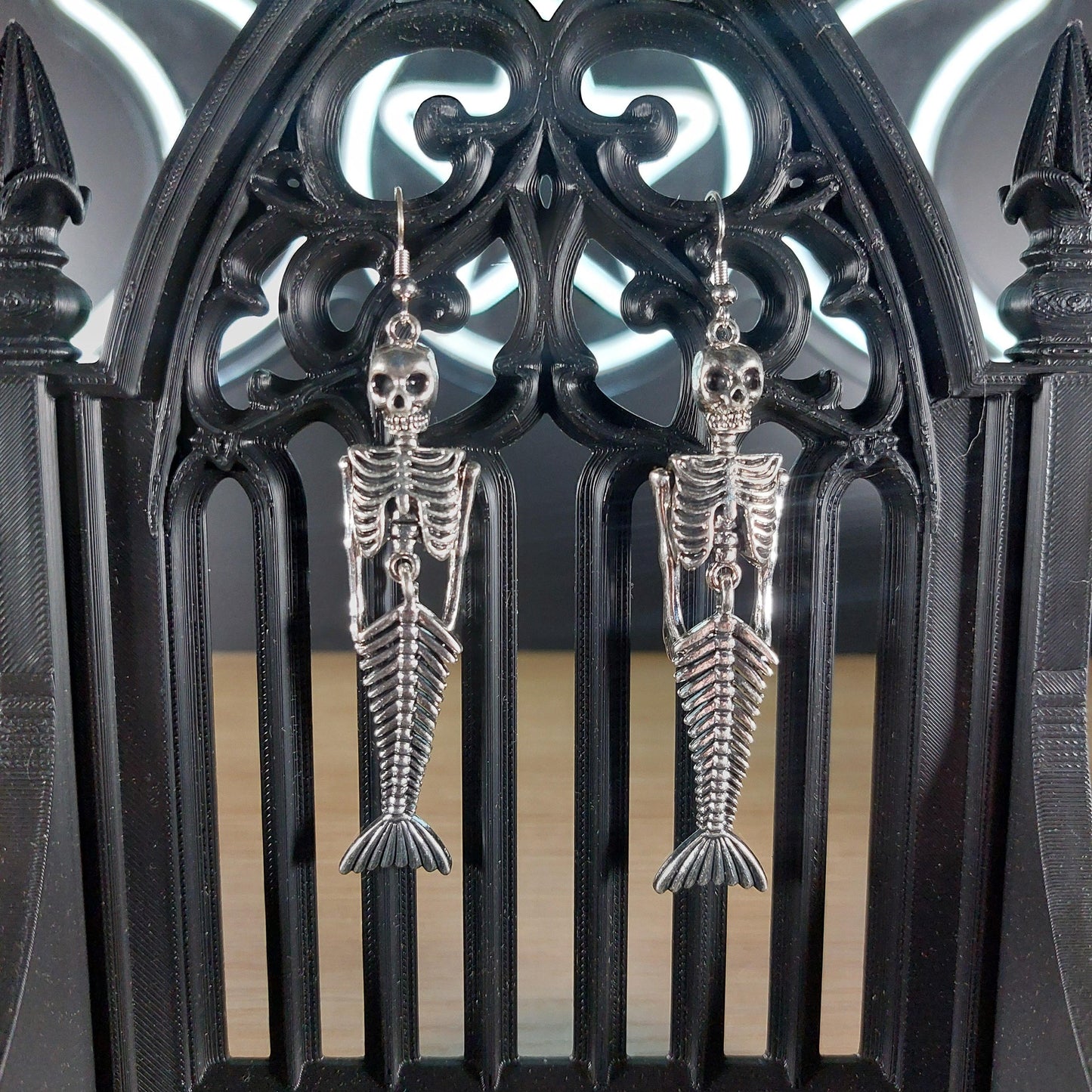 Silver tone mermaid skeleton dangle earrings hanging on a black gothic arch.