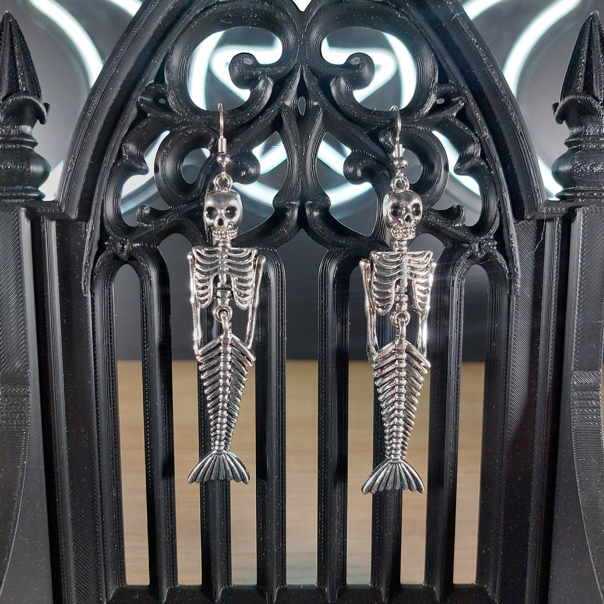 Silver tone mermaid skeleton dangle earrings hanging on a black gothic arch.