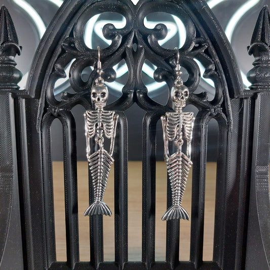 Silver tone mermaid skeleton dangle earrings hanging on a black gothic arch.