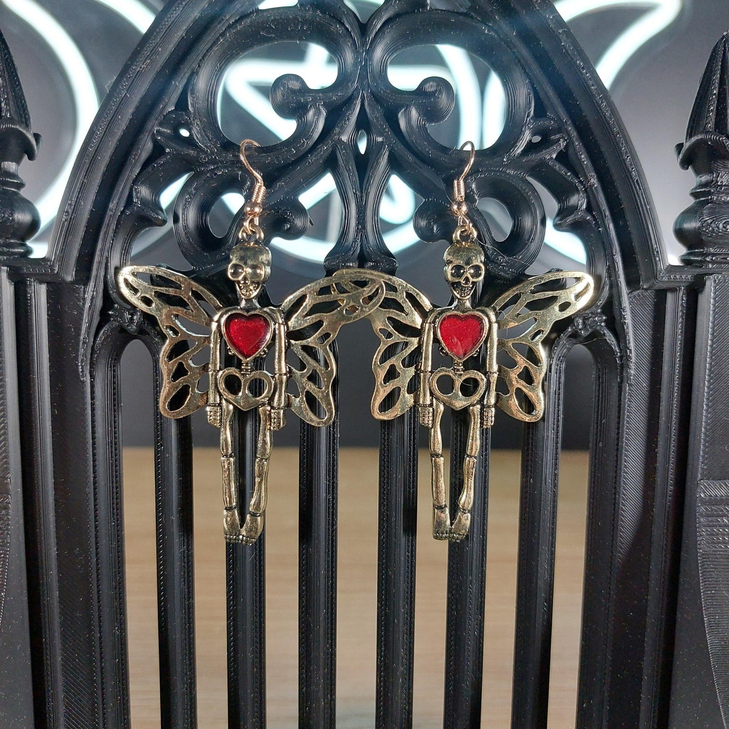 Gold tone skeleton with wings and red heart dangle earrings on a black gothic arch.