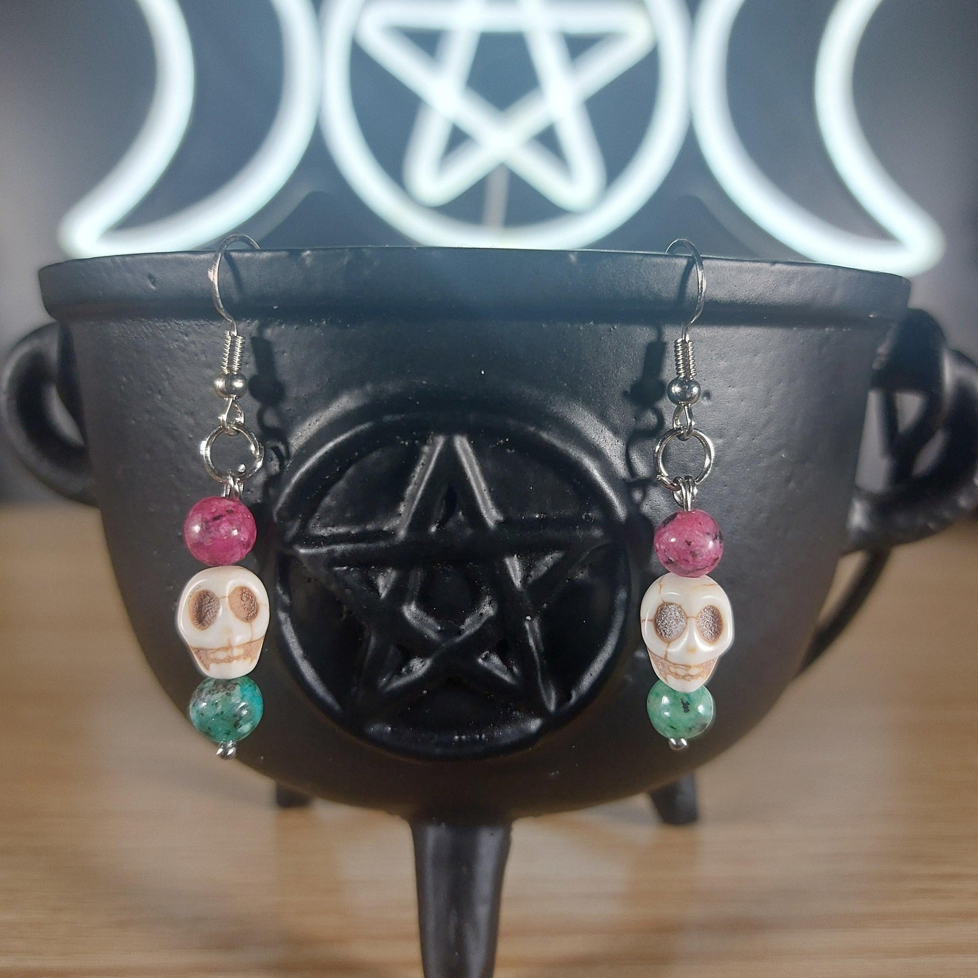 White howlite skull dangle earrings with pink and green glass beads on a black cauldron.