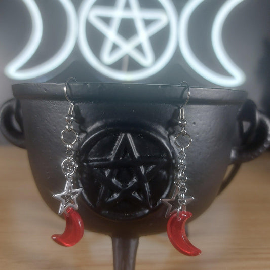 Red glass crescent moon and silver star dangle earrings on a black cauldron.