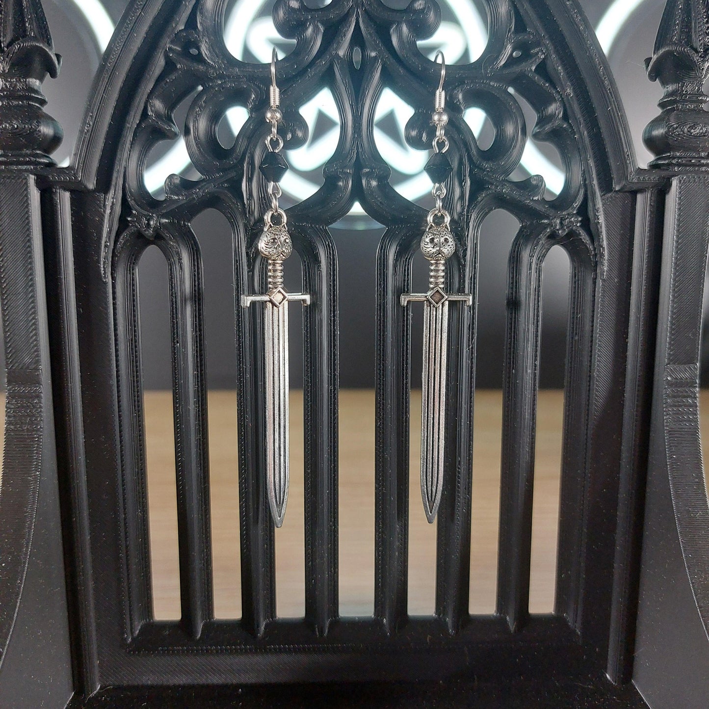 Silver tone sword dangle earrings with black bicone beads, hanging from a gothic black display.