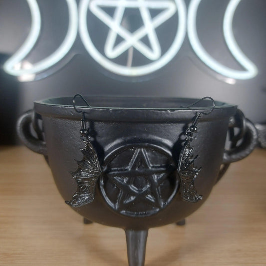 Black metal bat wing dangle earrings hanging from a black cauldron.