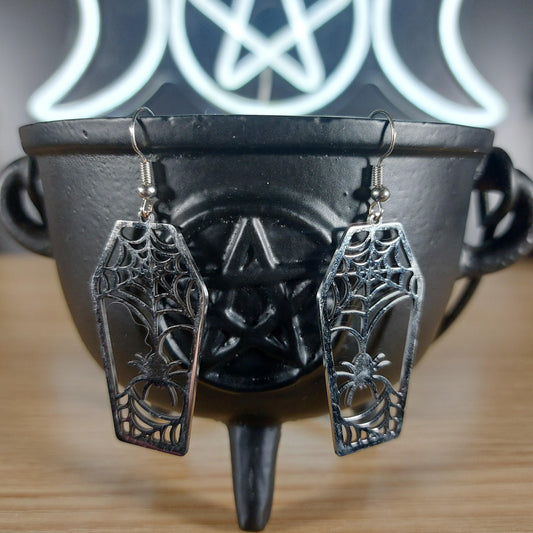 Silver-tone coffin dangle earrings with intricate spider web and spider cutouts, displayed on a black cauldron.